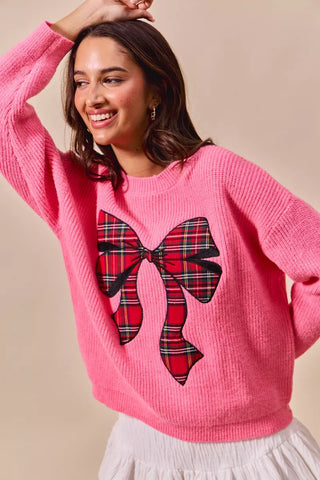 SO ME pink holiday sweater with a large plaid bow, shown with one sleeve raised.