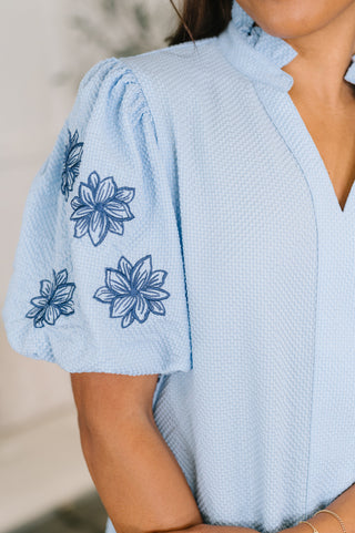 Light-blue That Easy blouse with floral-embroidered sleeves, close-up of the embroidered sleeve and ruffled collar.