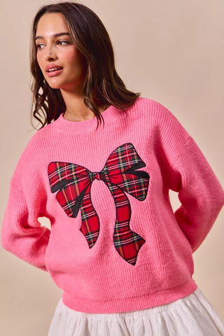Front view of the SO ME pink holiday sweater with a large red-and-black plaid bow design.
